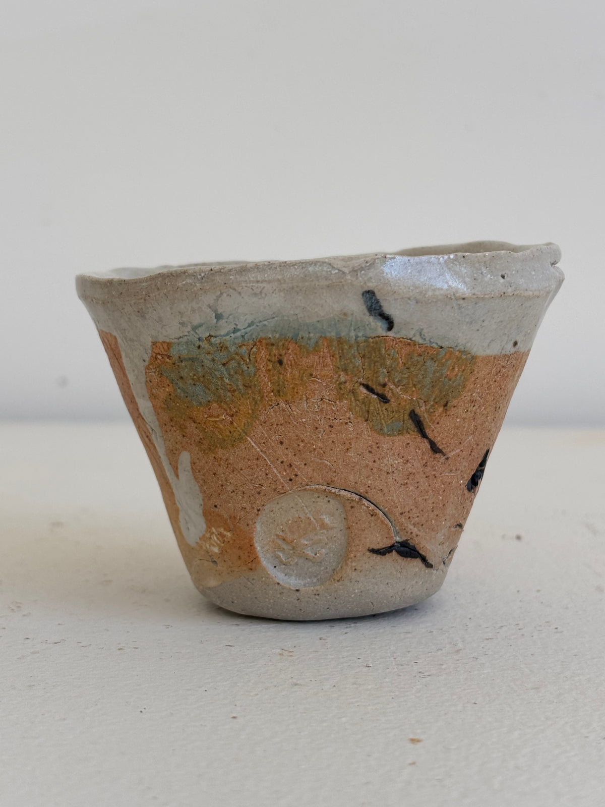 A handmade ceramic cup with a rustic, hand-painted finish. The cup features a terracotta orange-brown body with patches of light blue-green glaze, a white glazed rim, and small black speckles, set against a plain light gray background.
