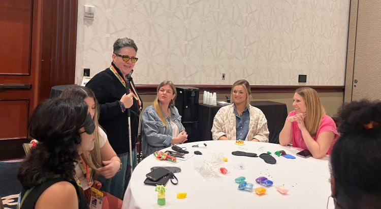 Cheri Owen speaks with a group of women at a round table during a workshop, holding a white cane and wearing yellow glasses.