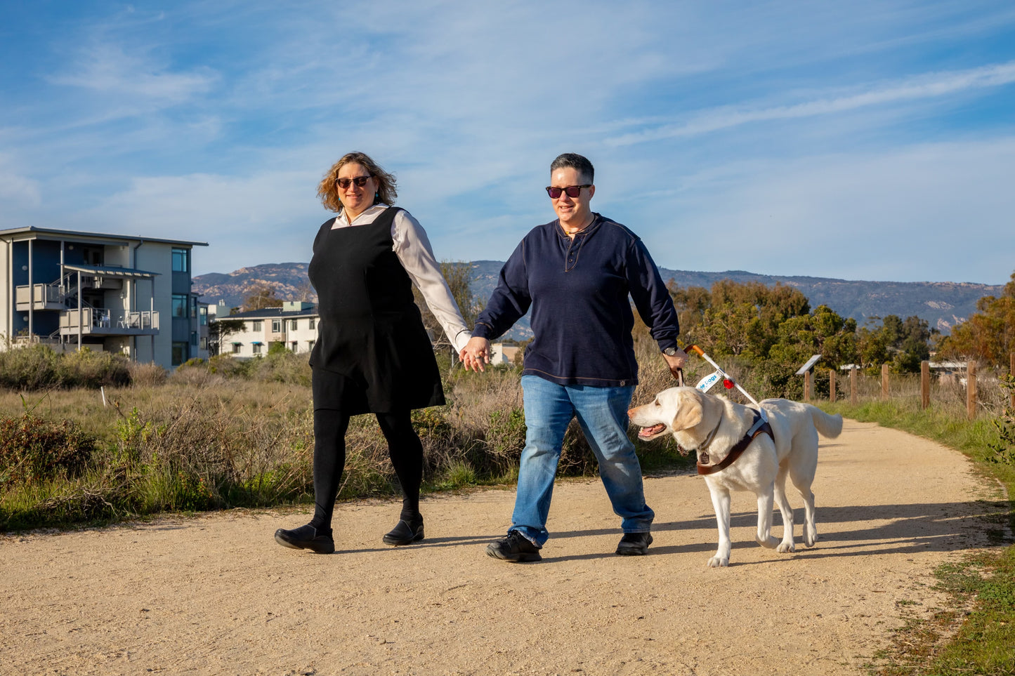 Cheri holds hands with wife Jenni while walking outdoors with Martinez, a guide dog, on a scenic path near homes.