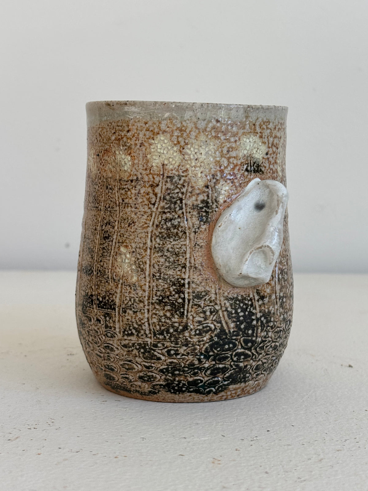 A handmade ceramic mug with a textured, speckled brown and black glaze, featuring a raised white ceramic bird detail on the side, set against a plain light background.