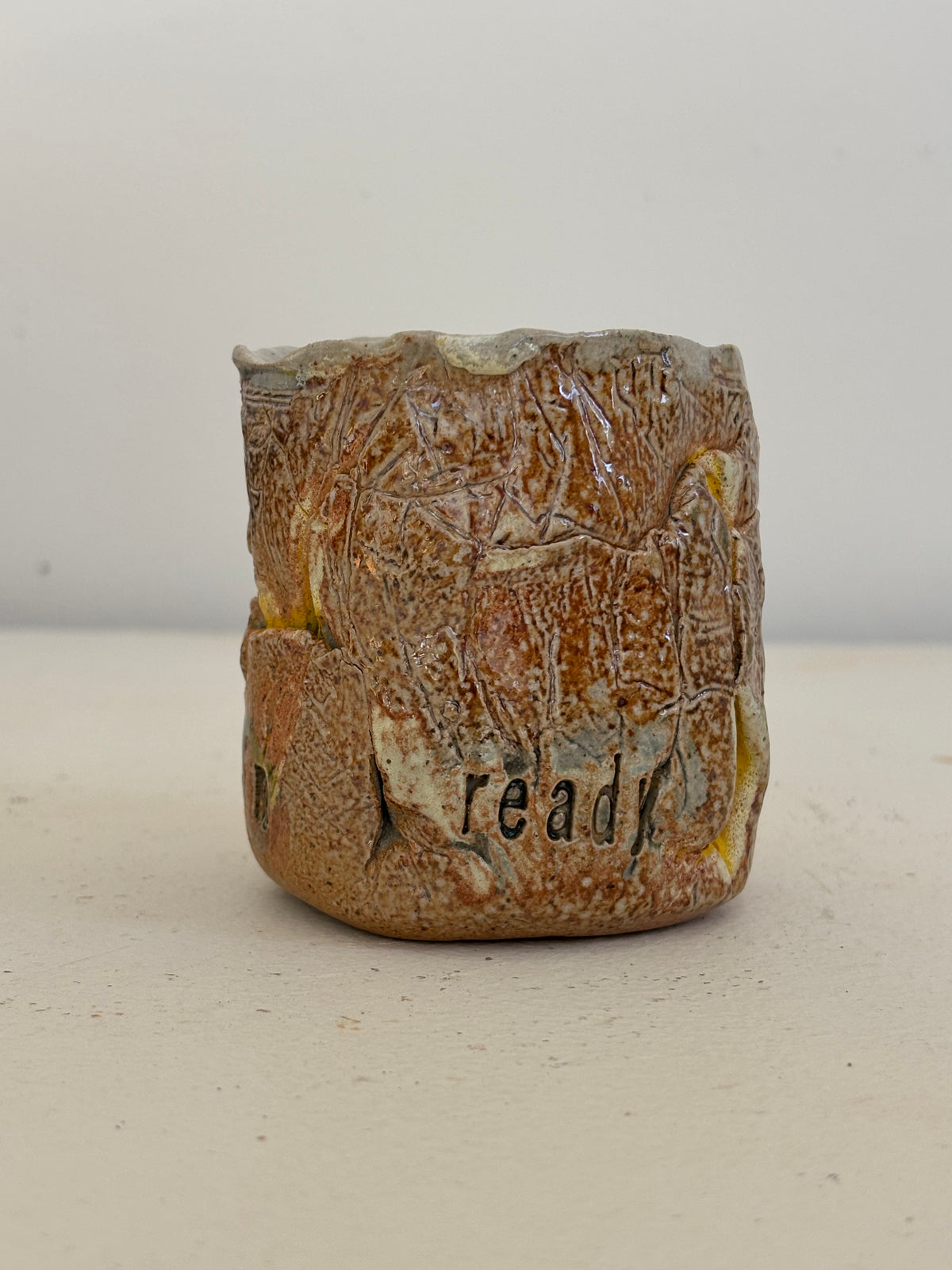 A handmade ceramic cup with a rustic, textured brown and beige glaze, featuring the word 'ready' hand-painted on the front, set against a plain light background.