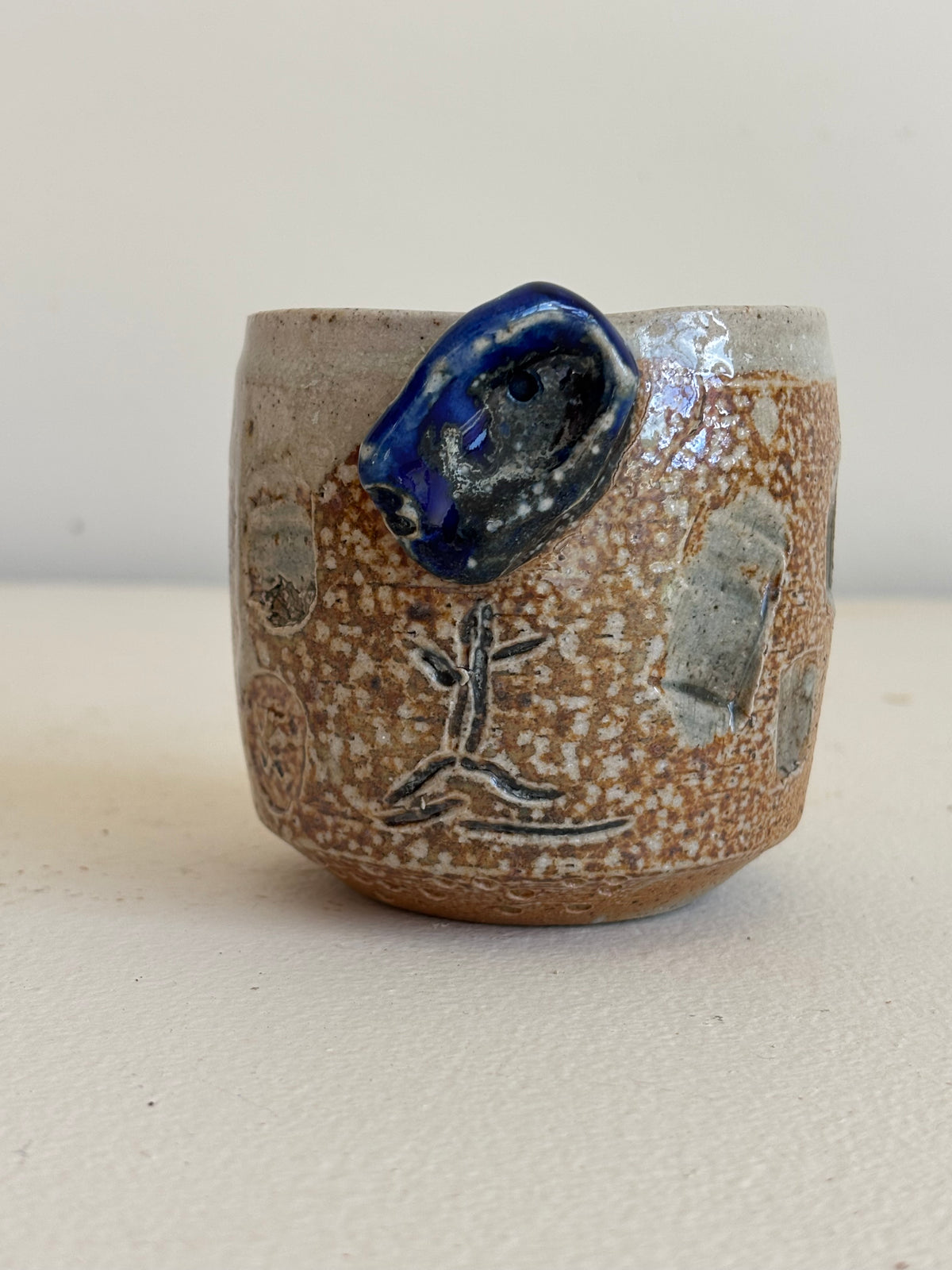 A handmade ceramic cup with a speckled brown and beige glaze, featuring a protruding dark blue glazed protrusion and a simple black painted figure on the front, set against a plain light background.