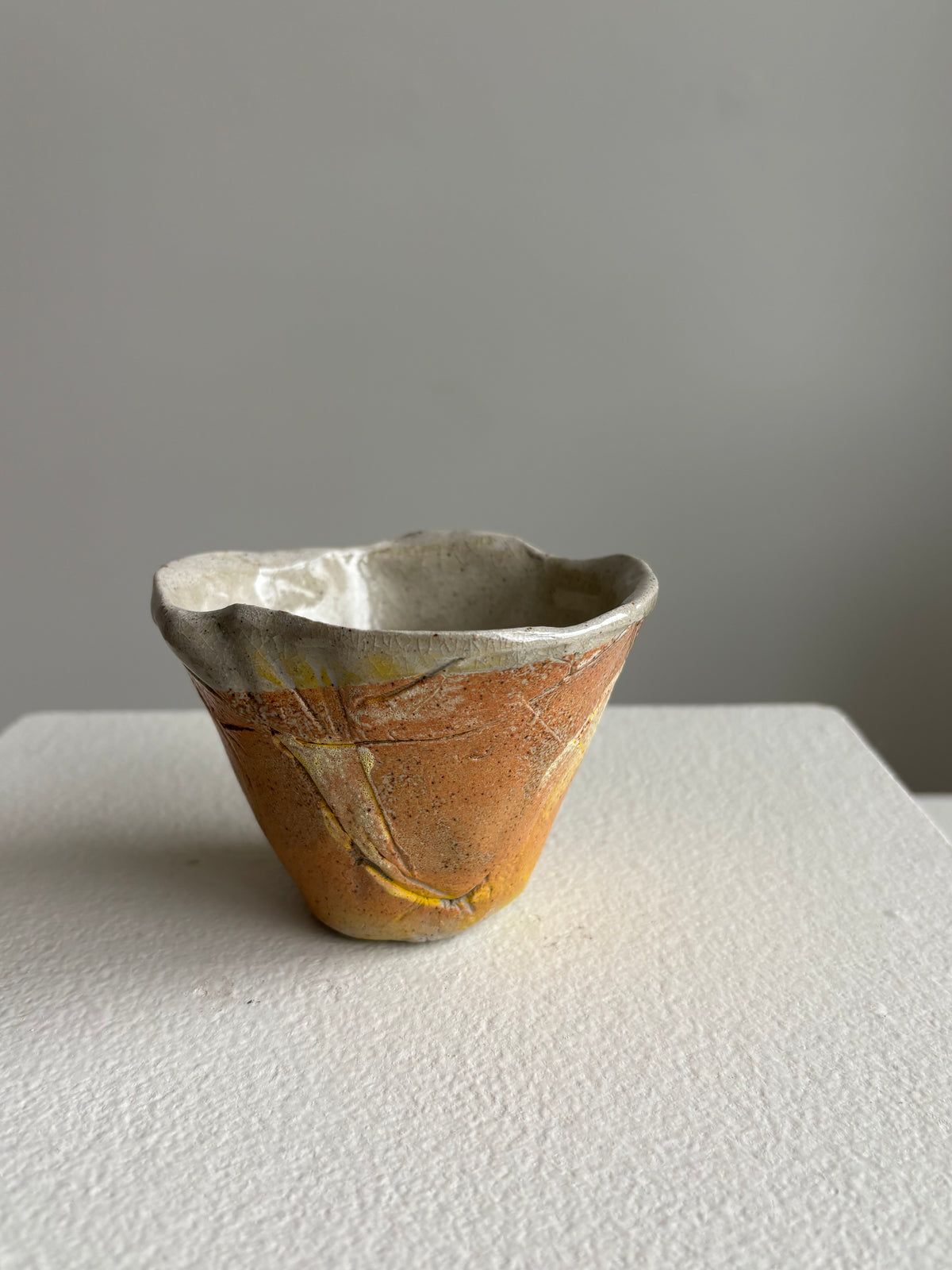 A small handmade ceramic cup with a textured, crackled glaze in earthy orange-brown, cream, and yellow tones, sitting on a white textured surface against a neutral gray background.AC