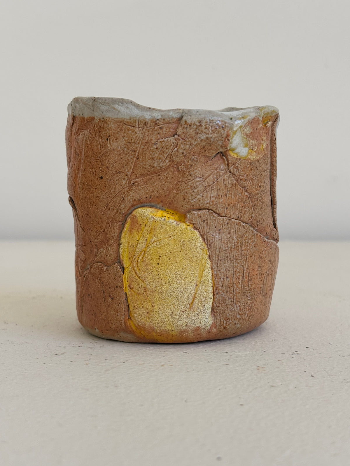 A handmade ceramic cup with a rustic, earthy brown and beige textured glaze, featuring a prominent metallic gold leaf inlay on the front and a light gray glazed rim.