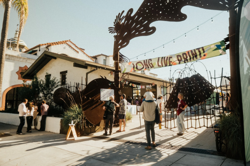 Outdoor festival scene with people walking under a large decorative tree sculpture and a banner that reads "LONG LIVE POINT!" in front of a white building with palm trees.