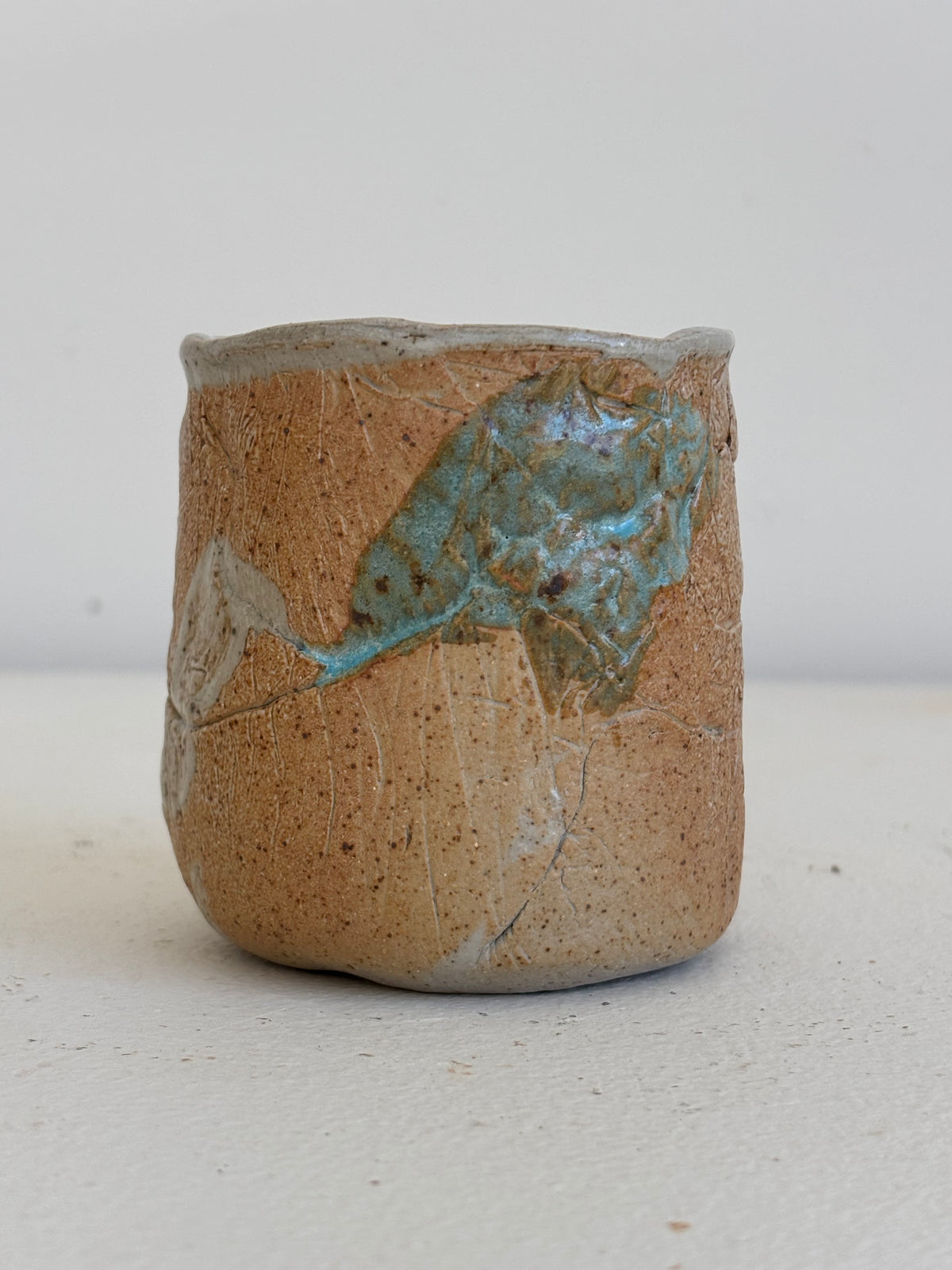 A handmade ceramic cup with a textured, speckled tan-brown glaze and a prominent iridescent turquoise-blue patch on the side, set against a plain light background.