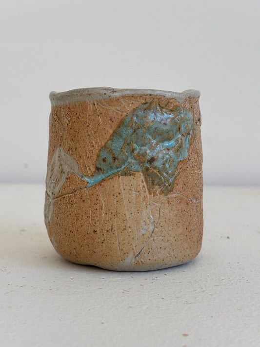 A handmade ceramic cup with a textured, speckled tan-brown glaze and a prominent iridescent turquoise-blue patch on the side, set against a plain light background.
