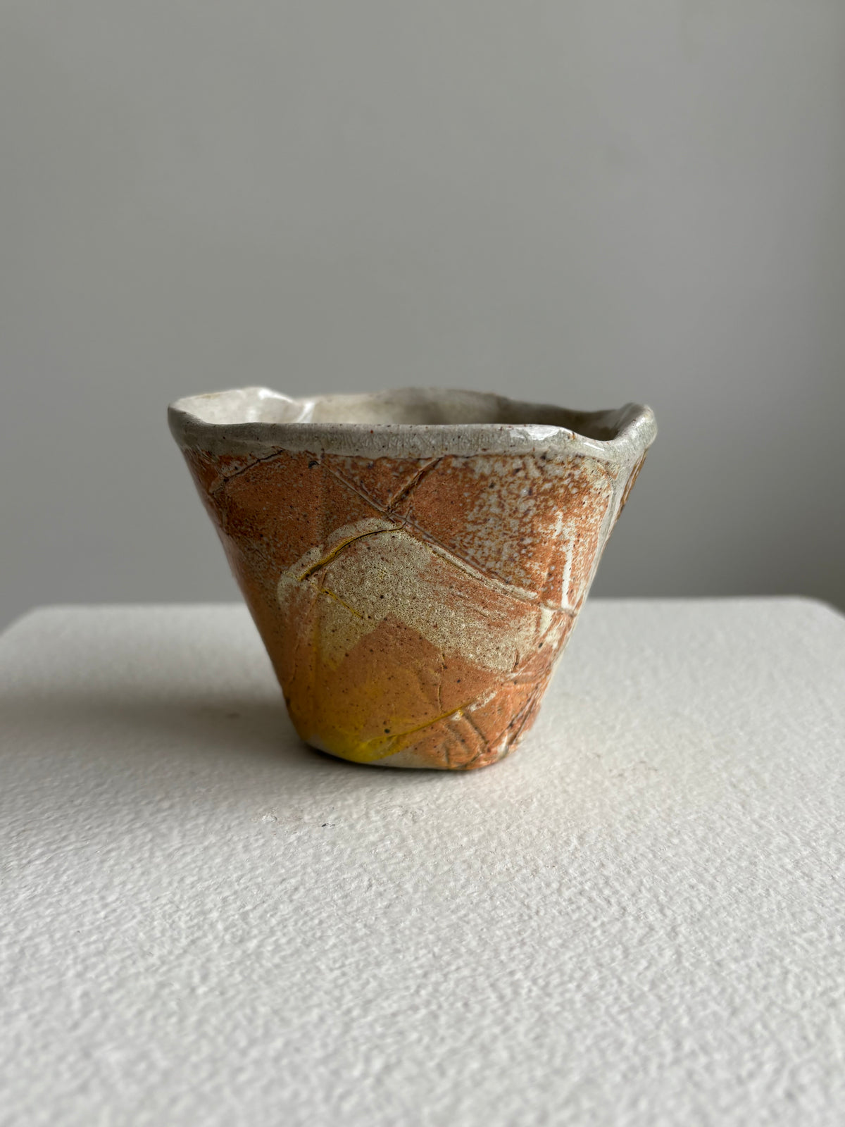A small handmade ceramic cup with a textured, crackled glaze in earthy orange-brown, cream, and yellow tones, sitting on a white textured surface against a neutral gray background.AC