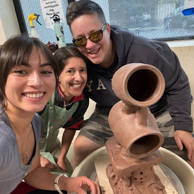 Cheri, Mia, and a student smile together behind a large clay sculpture on a pottery wheel in the studio.