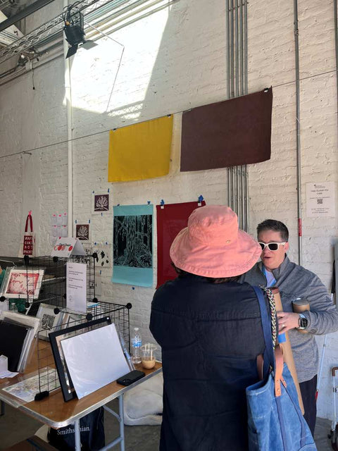 Cheri Owen talking with visitors at her booth, featuring framed prints and large artwork hanging on a brick wall.