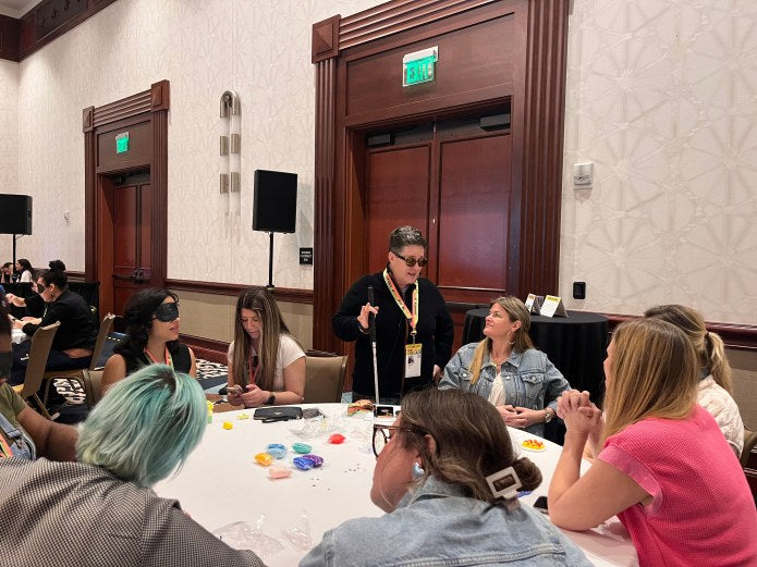 Cheri Owen stands with a white cane, speaking to a group of participants seated around a table with tactile art supplies.