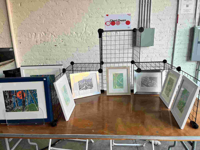 Framed prints displayed on a folding grid setup with a Cheri Owen nameplate at a table inside the Off Register event.