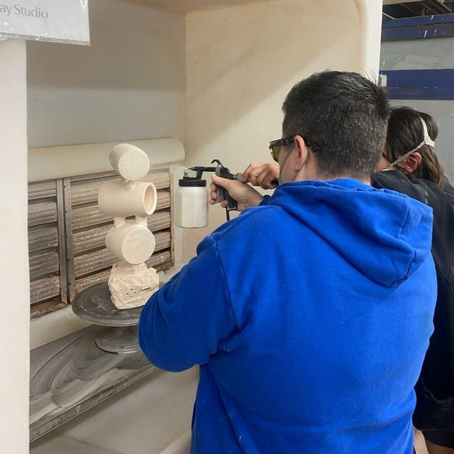 Cheri uses a spray gun to glaze a stacked pottery sculpture in a spray booth, wearing safety glasses and a blue hoodie.