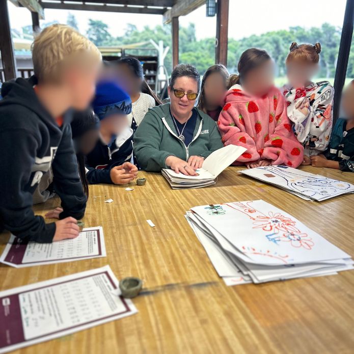 Cheri sits at a table surrounded by children, showing tactile art books; children's faces are blurred for privacy.