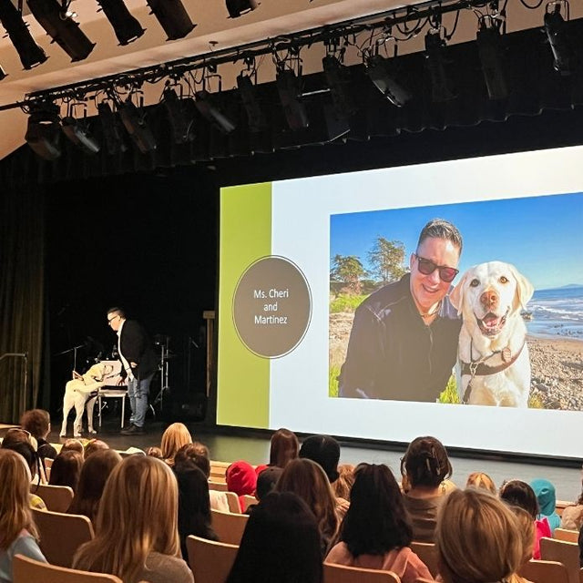 Cheri and her guide dog Martinez stand on stage presenting to a student audience, with a large slide of their beach photo displayed behind them.