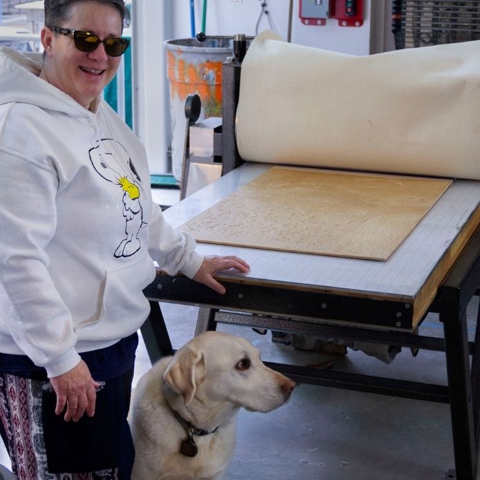 Cheri stands next to her guide dog Martinez by a large printing press, smiling while a printmaking plate is set up on the press.