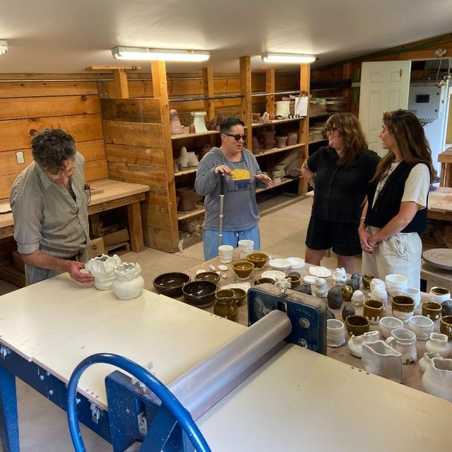 Cheri talks with three visitors in a ceramics studio, surrounded by shelves and tables filled with pottery pieces and tools.