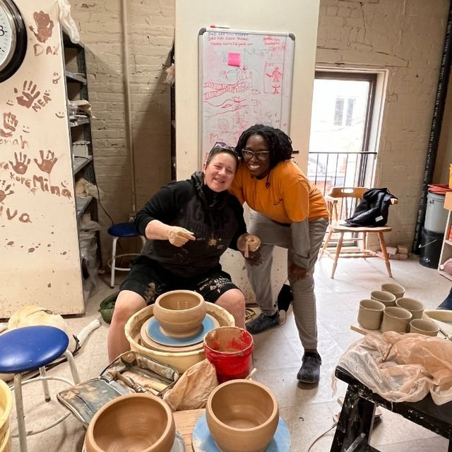 Cheri sits at a pottery wheel shaping a clay bowl while smiling with a friend beside her in a ceramics studio.