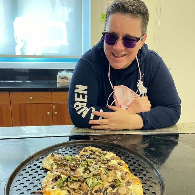 Cheri smiling and leaning on a counter beside her freshly baked pizza topped with olives, mushrooms, and greens.