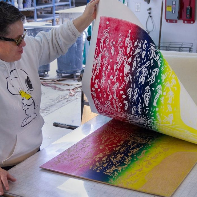 Cheri lifting a vivid rainbow print from a press table, revealing intricate plant-inspired patterns.