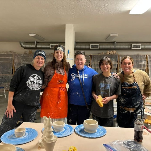 Cheri stands with four students in a ceramics studio, all smiling with pottery pieces on the table in front of them.