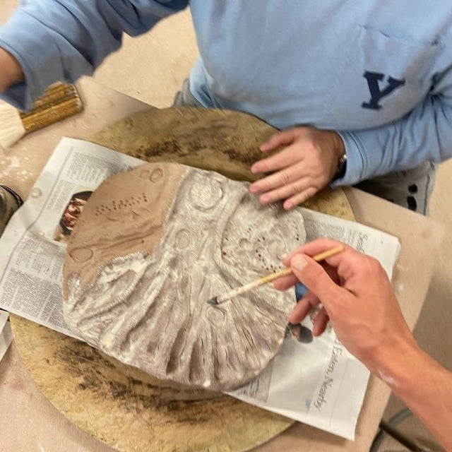 Close-up of hands working on a textured pottery piece at the studio, with Cheri wearing a blue Yale sweatshirt.