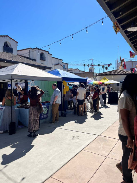 Visitors explore artist booths under white tents in a sunny courtyard at the Santa Barbara print fair event.