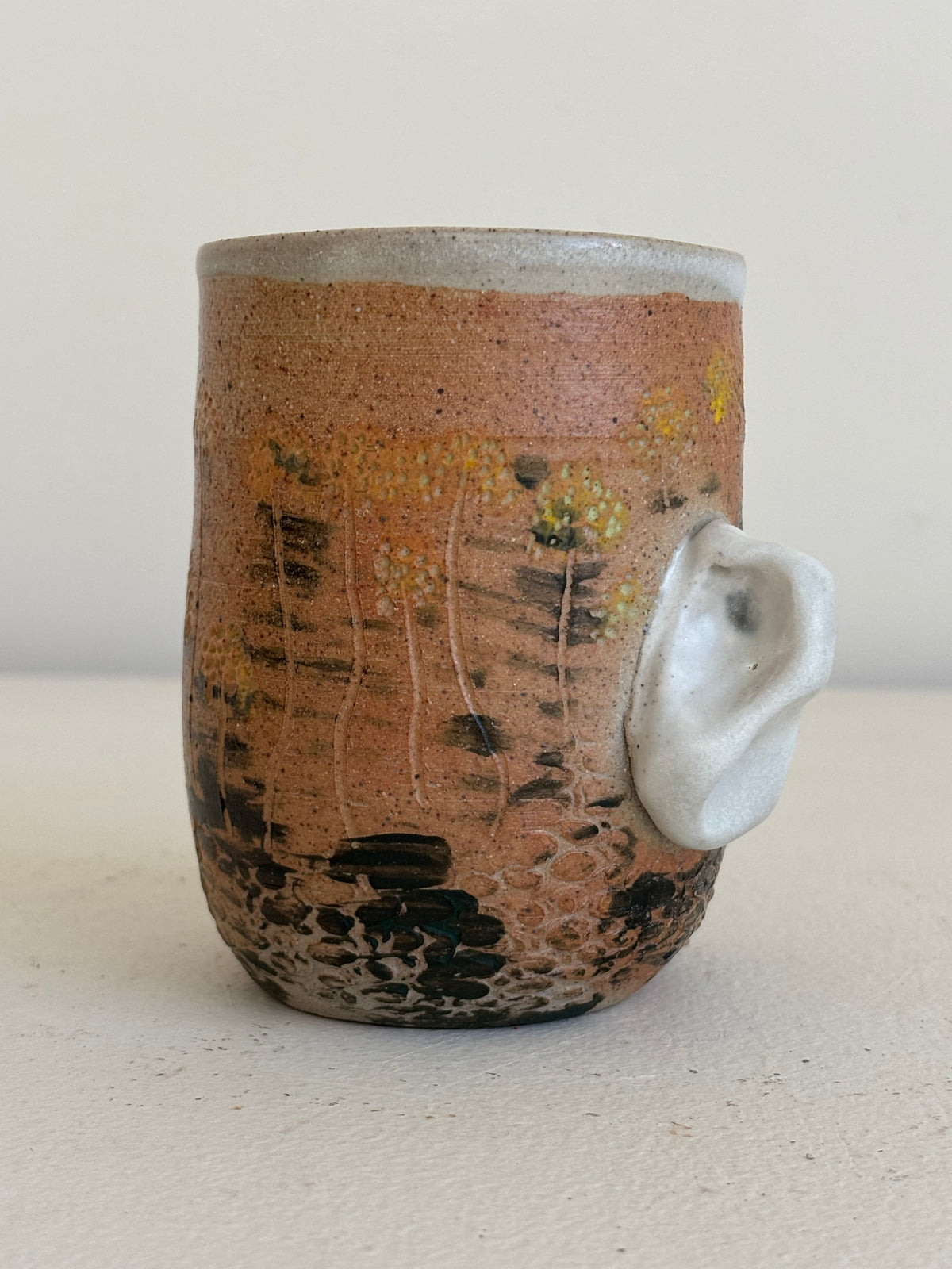 A handmade ceramic mug with a rustic terracotta-brown glaze, hand-painted wildflower patterns, and a sculptural white animal ear handle protruding from the side, set against a neutral beige background.