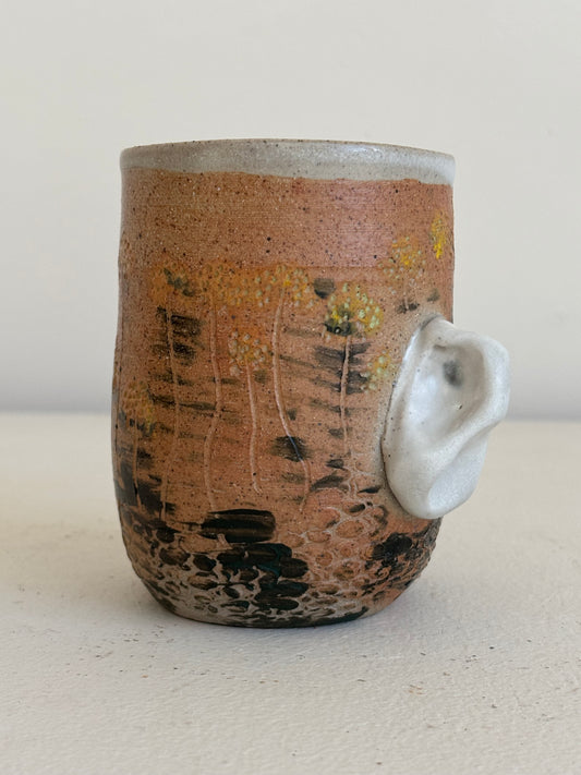 A handmade ceramic mug with a rustic terracotta-brown glaze, hand-painted wildflower patterns, and a sculptural white animal ear handle protruding from the side, set against a neutral beige background.