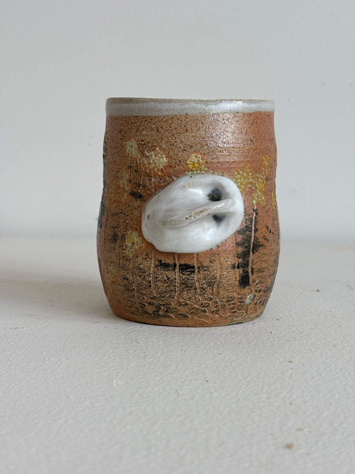 A small handmade ceramic cup with a textured brown and orange glaze, featuring a protruding white shell-like ornament on the front, set against a plain light gray background.