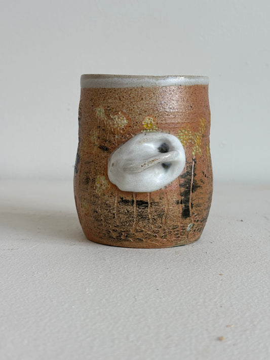 A small handmade ceramic cup with a textured brown and orange glaze, featuring a protruding white shell-like ornament on the front, set against a plain light gray background.