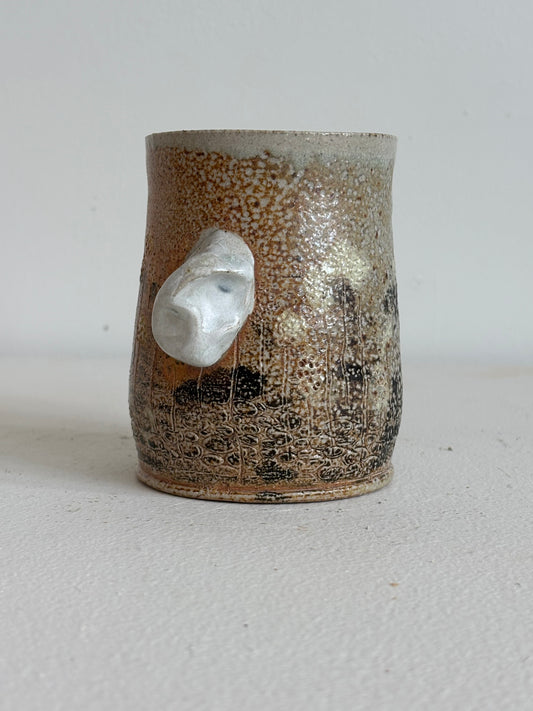 A handmade ceramic mug with a textured, speckled brown and gold glaze, featuring a unique organic white protruding side detail, set against a plain light gray background.
