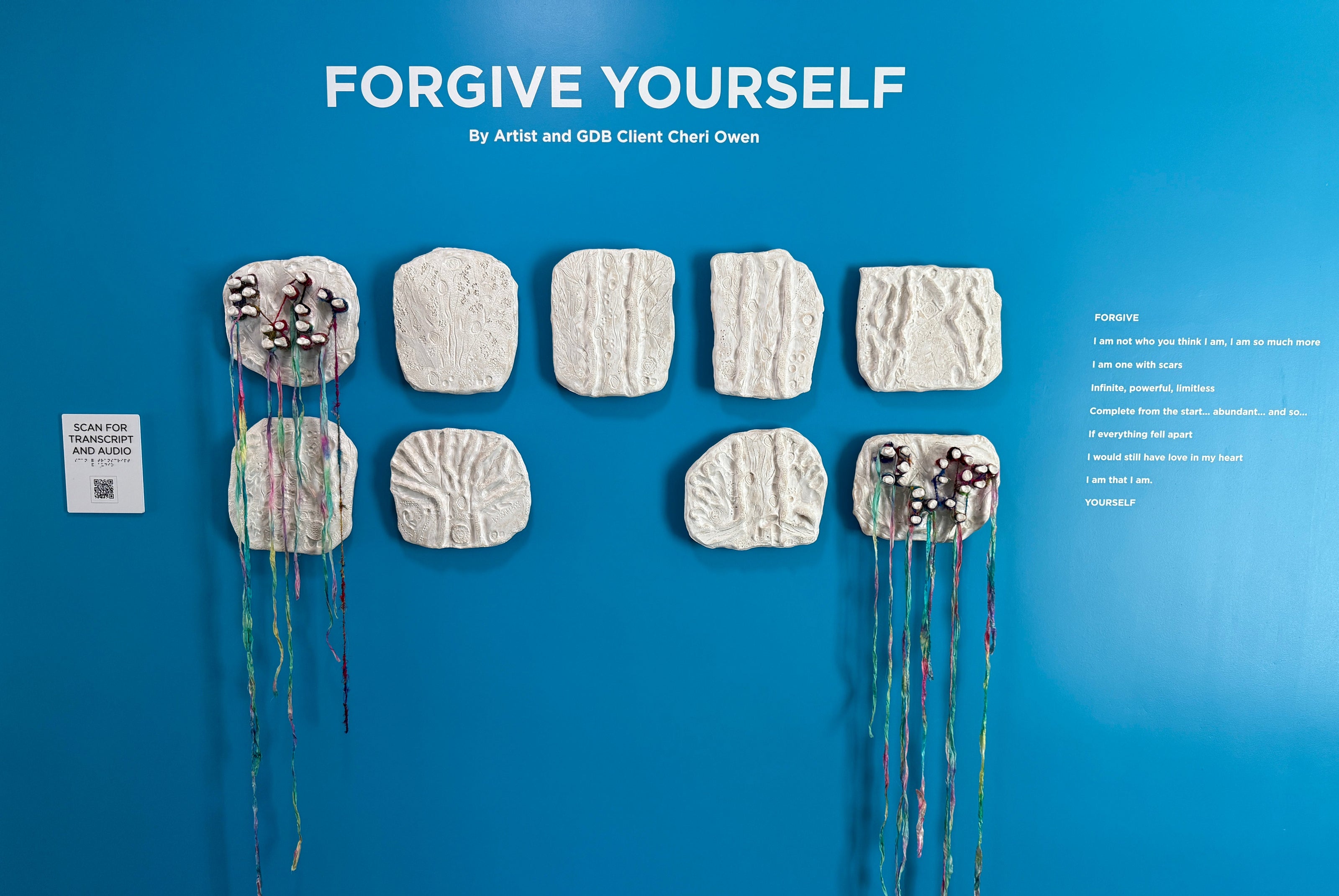 Installation titled "Forgive Yourself" by artist Cheri Owen, featuring twelve white ceramic tiles arranged in a grid on a bright blue wall. Some tiles have textured imprints resembling nature, while three include colorful yarn and tactile elements. A poem and a QR code for transcript and audio are also displayed on the wall.