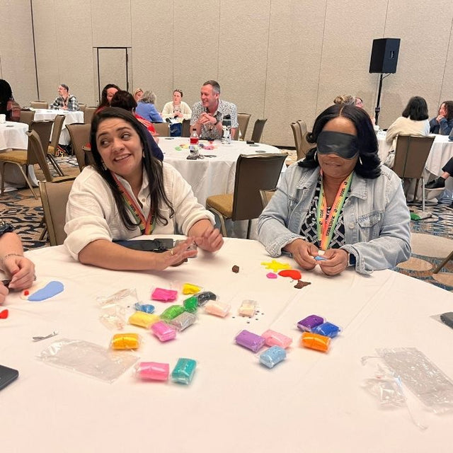 Two women participate in a tactile clay workshop; one wears a blindfold while shaping colorful clay.