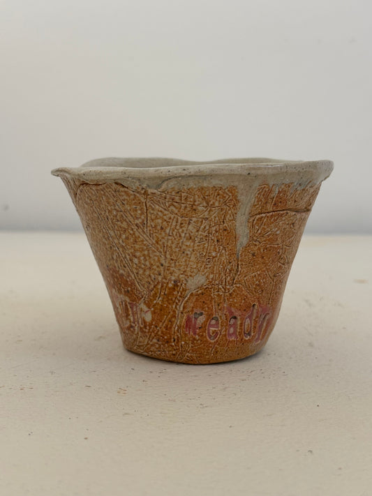 A handmade ceramic cup with a textured, crackled orange-brown glaze and a light beige rim, featuring the word 'ready' in pink lettering on the side, set against a plain off-white background.