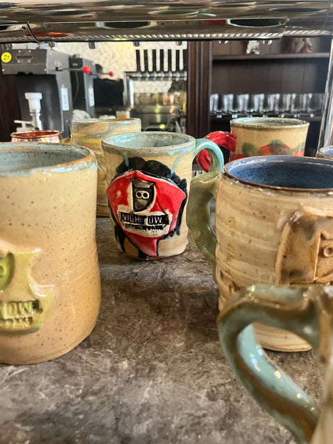 Handmade ceramic mugs with Night Owl logo displayed on counter at event.
