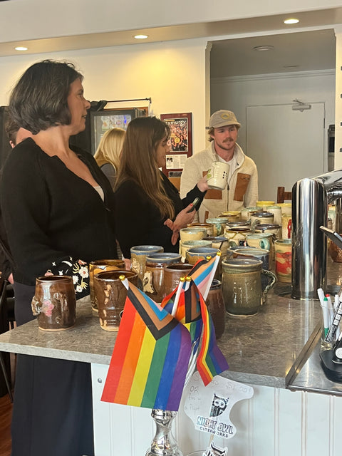 Guests in conversation around counter with ceramic mugs and Pride flag at Night Owl Opening