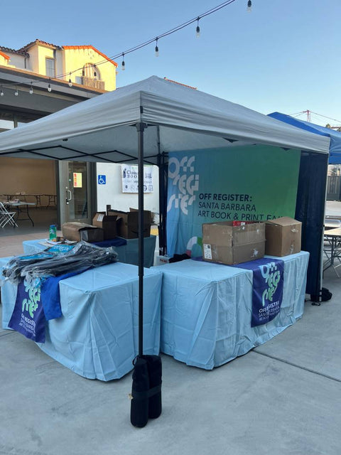 Outdoor booth with Off Register banner, blue table covers, and event merchandise at the Santa Barbara print fair.
