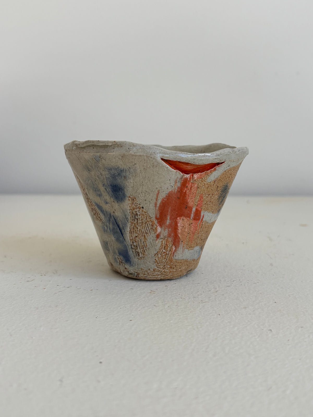 A handmade ceramic cup with a rustic, hand-painted glaze featuring blue, orange, and brown brushstrokes on a light beige base, set against a plain light gray background.
