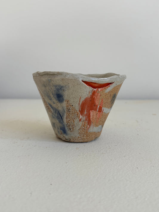 A handmade ceramic cup with a rustic, hand-painted glaze featuring blue, orange, and brown brushstrokes on a light beige base, set against a plain light gray background.