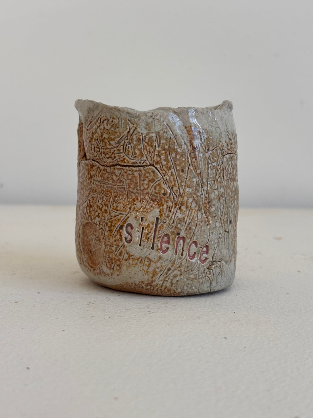 A small handmade ceramic cup with a textured, earthy brown and beige glaze, featuring the word 'silence' printed in red on the front, set against a plain light background.