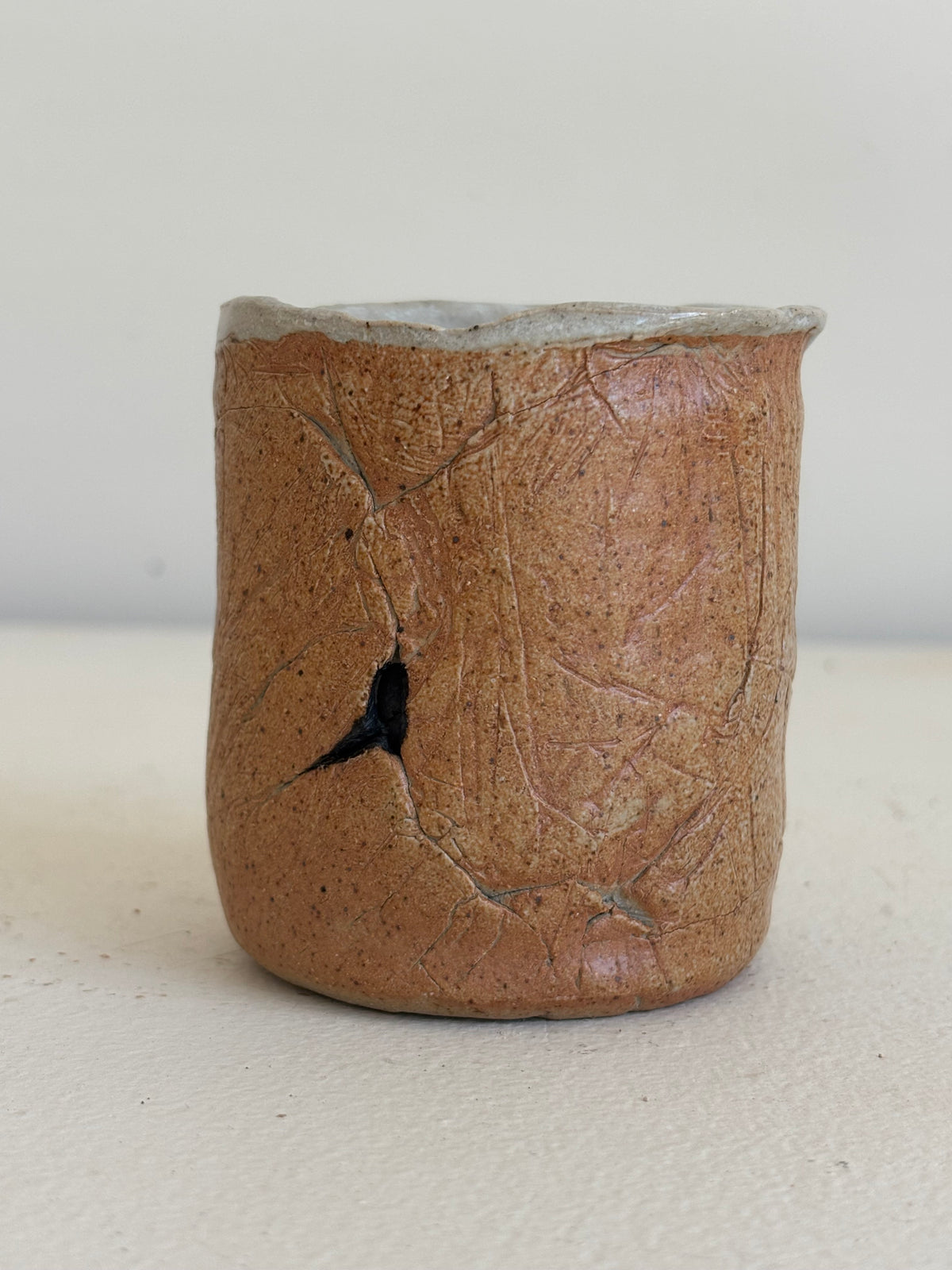 A handmade ceramic cup with a textured, earthy brown glaze and a visible crack on the front, set against a plain light background.