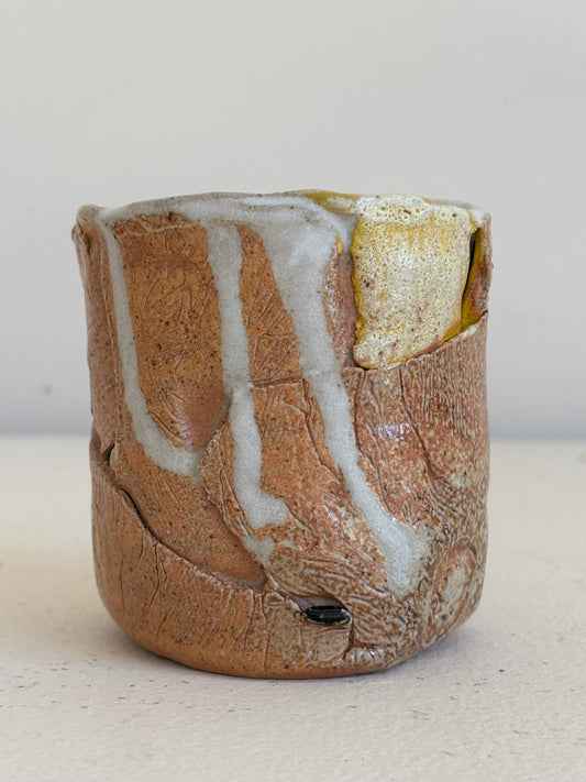 A handmade ceramic cup with a textured, earthy brown body and white glaze drips, featuring a small yellow accent on the upper right side, set against a plain light background.