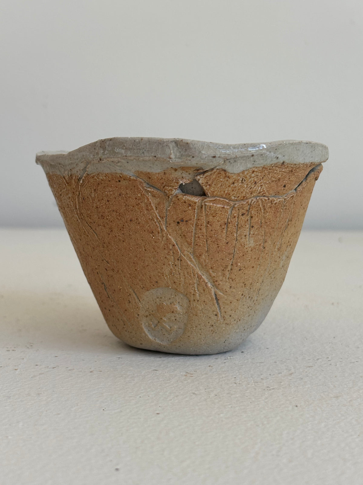 A handmade ceramic cup with a rustic, hand-rolled clay texture in earthy brown and beige tones, featuring a slightly lighter gray-beige glazed rim and a small dark crack on the front.