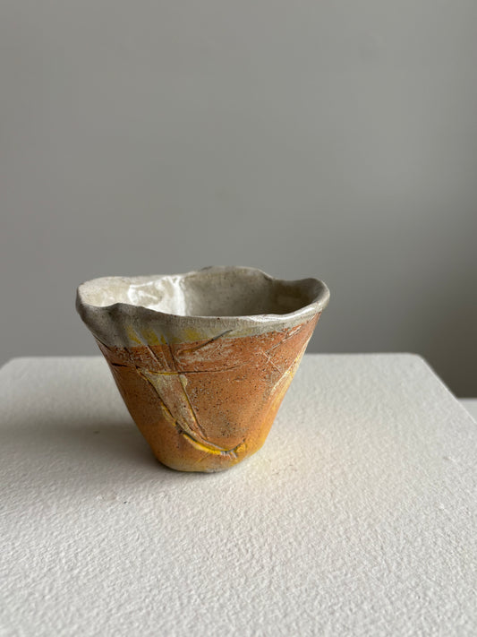 A small handmade ceramic cup with a textured, crackled glaze in earthy orange-brown, cream, and yellow tones, sitting on a white textured surface against a neutral gray background.AC