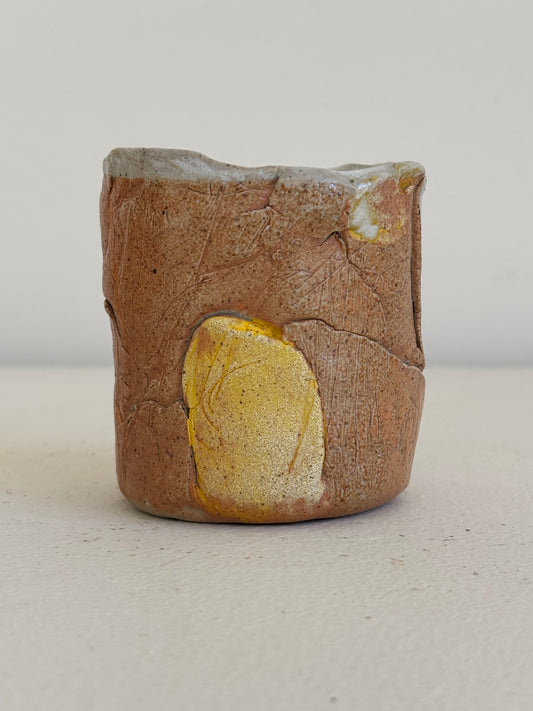 A handmade ceramic cup with a rustic, earthy brown and beige textured glaze, featuring a prominent metallic gold leaf inlay on the front and a light gray glazed rim.
